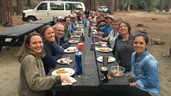 A large group of people enjoying a meal at a long picnic table in a wooded area