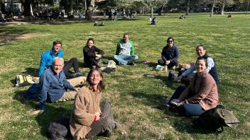 A group of eight people sitting in a circle on grassy parkland.