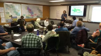 A group of people in a meeting room with maps on the walls, viewing a presentation on a "Ecosystem Service Workshop"