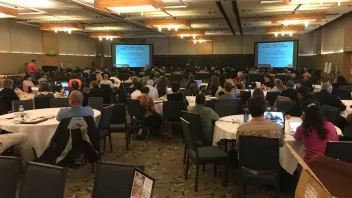 A conference room filled with attendees facing two projection screens.
