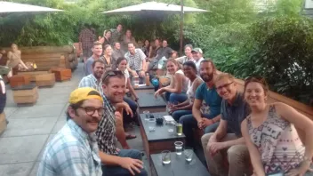 A large group of people seated at an outdoor patio under umbrellas, surrounded by green foliage.