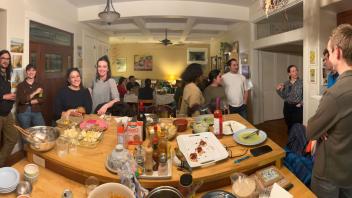 A social gathering in a kitchen with people chatting and food laid out on a countertop.