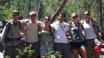 A group of six people smiling in a forest, dressed for hiking.