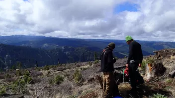 Two people stand on a rocky ridge overlooking forested mountains under a cloudy sky.