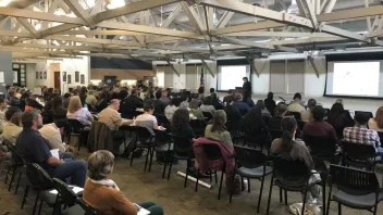 A large group of people seated in a conference room attending a presentation with two projection screens.