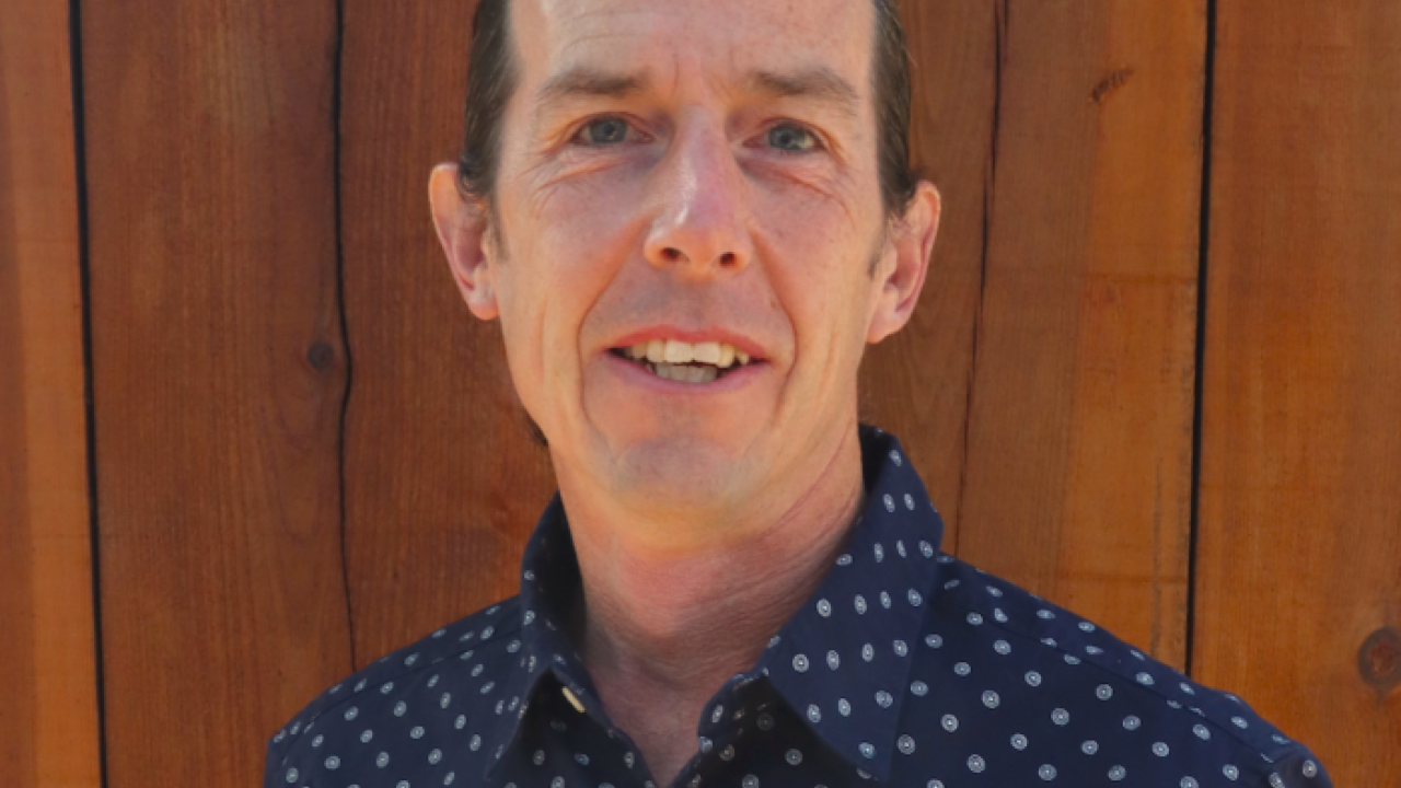Person wearing a dark blue patterned shirt in front of a wooden background.