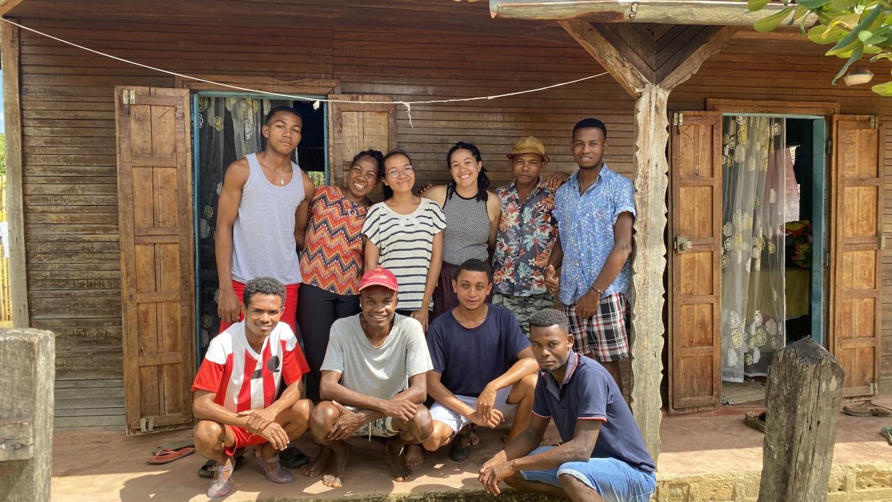 A group of ten people in front of a wooden house with open doors and floral curtains.
