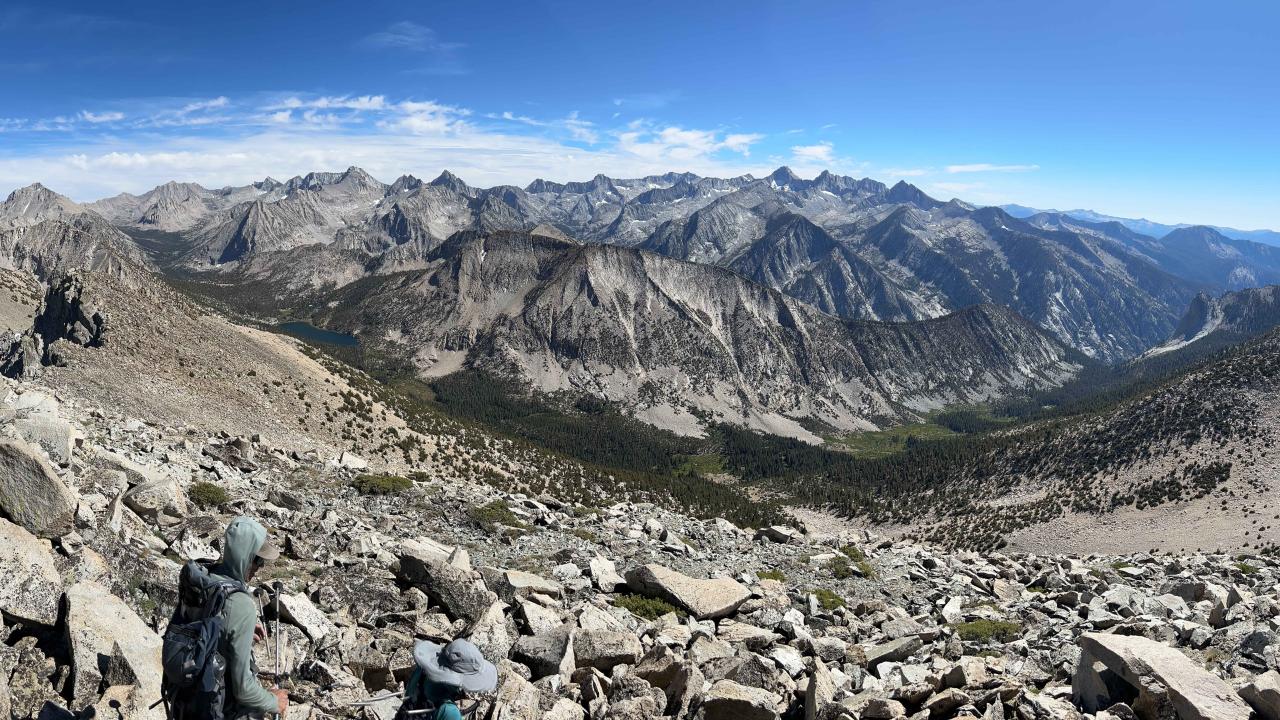 A panoramic view of a mountainous landscape with rocky terrain, two hikers in the foreground, and distant peaks under a blue sky