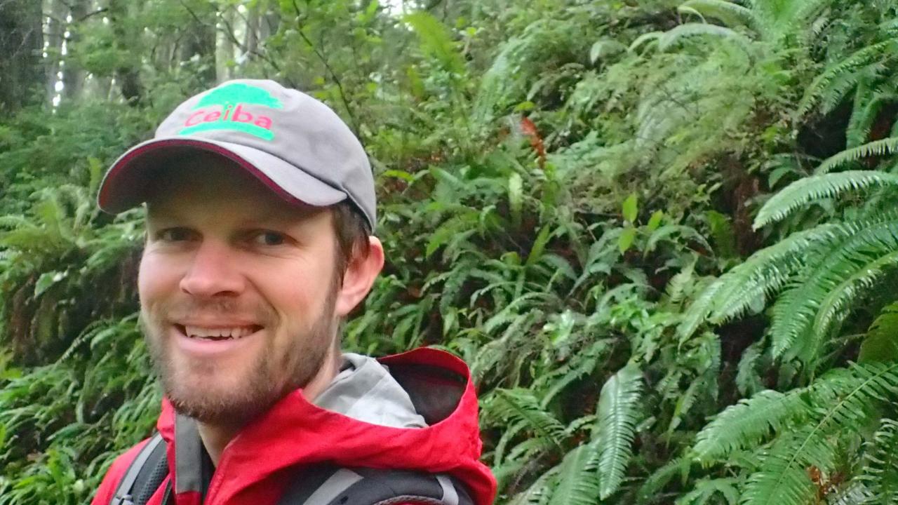 A man in a red jacket and gray cap stands in a green forest