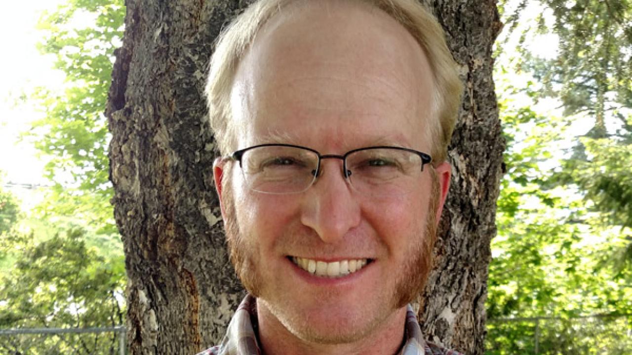 Smiling man in glasses with sideburns standing in front of a tree