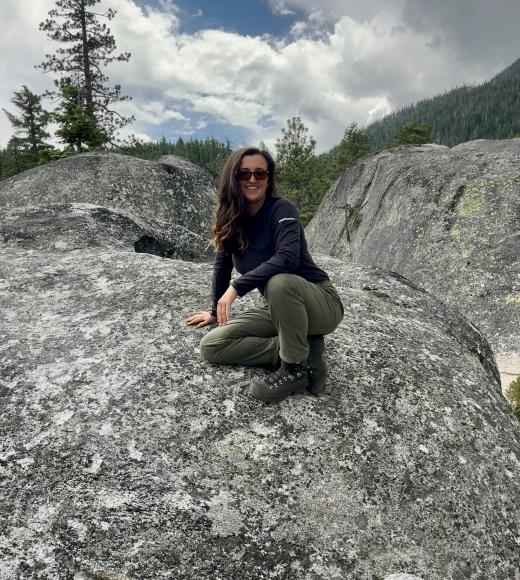 Person crouching on a large gray rock, surrounded by trees and hills under a cloudy sky.