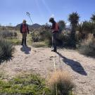 Two individuals in safety gear measure land in a desert landscape under a clear sky.