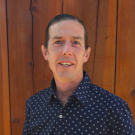 Person wearing a dark blue patterned shirt in front of a wooden background.