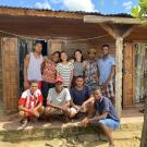 A group of ten people in front of a wooden house with open doors and floral curtains.