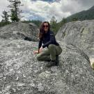 Person crouching on a large gray rock, surrounded by trees and hills under a cloudy sky.