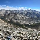 A panoramic view of a mountainous landscape with rocky terrain, two hikers in the foreground, and distant peaks under a blue sky