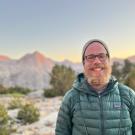 Person smiling outdoors with mountains at sunset in the background.
