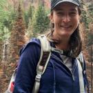 Person in outdoor attire with a mountain forest backdrop.