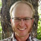 Smiling man in glasses with sideburns standing in front of a tree