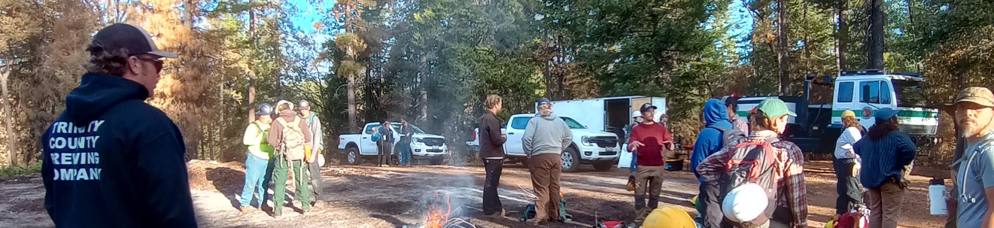 People at a forest fire training session with a small fire in a forest clearing.